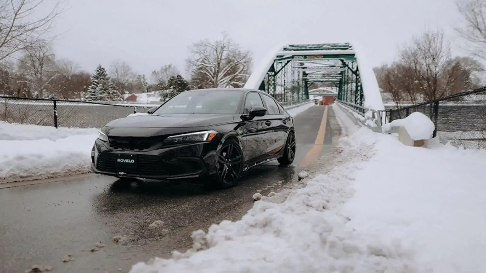 Black sedan equipped with winter tires parked on snowy road near a green bridge in winter landscape.