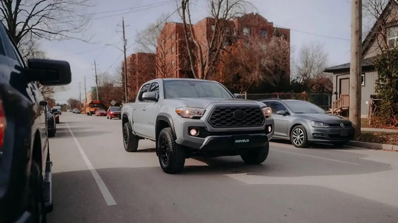 Gray pickup truck with all terrain tires driving on urban street surrounded by residential buildings and other vehicles.