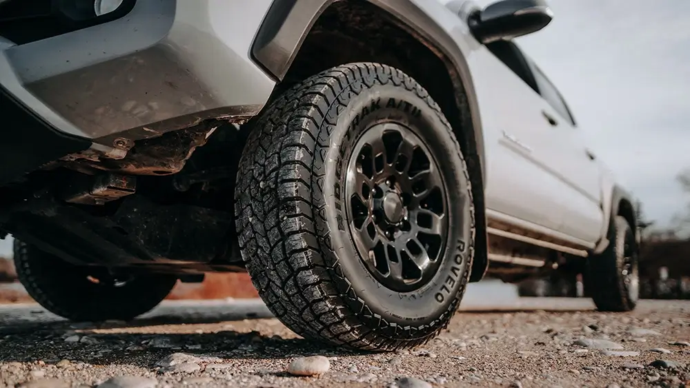 All terrain tires on a truck, showcasing detailed tread pattern, parked on gravel road in outdoor setting.