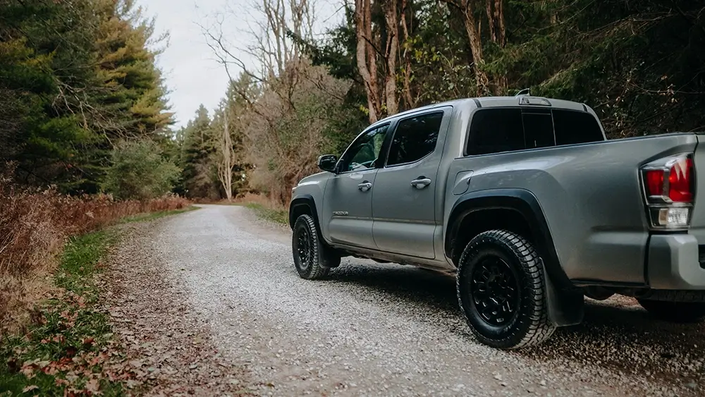 Tacoma truck with all terrain tires parked on gravel forest road surrounded by trees